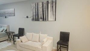 A living room with a beige sofa, a small round table, a dark wooden chair, and a painting of trees on the wall. - Cozy 1-bedroom studio apartment in perfect Houston with fitness room and AC, (Houston)