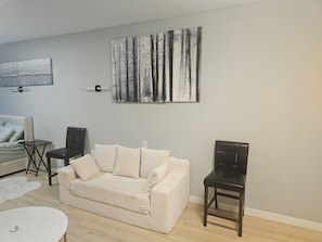 Uma sala de estar com sofá bege, uma mesinha redonda, uma cadeira de madeira escura e um quadro de árvores na parede.