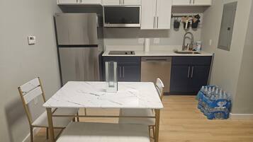 A compact kitchen with white cabinets, a stainless steel refrigerator, a dishwasher, and a dining area with a marble table and chairs.
