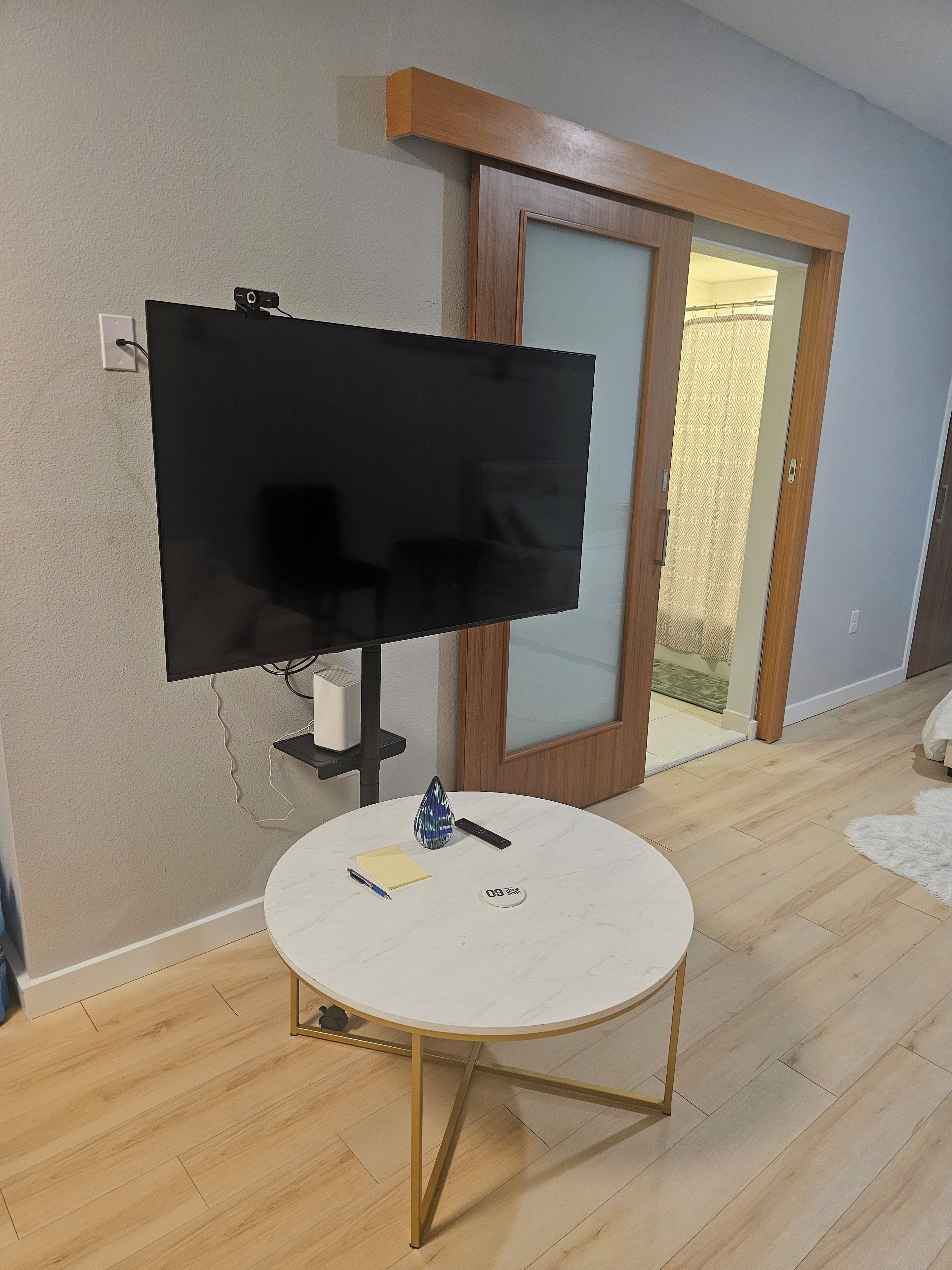 A room with a flat-screen TV mounted on the wall, a small round table with a pen, a remote, and a decorative item.