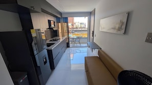 Et kompakt køkken med moderne hårde hvidevarer, et spiseområde med en bænk og udsigt til balkonen.