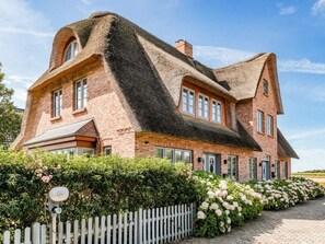 Ein großes Backsteinhaus mit einem steilen Dach, zahlreichen Fenstern und einem weißen Zaun aus Holzlatten.