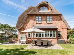 Ein Haus aus Backstein mit einem Strohdach, einer Terrasse mit Gartenmöbeln und einem großen Fenster.