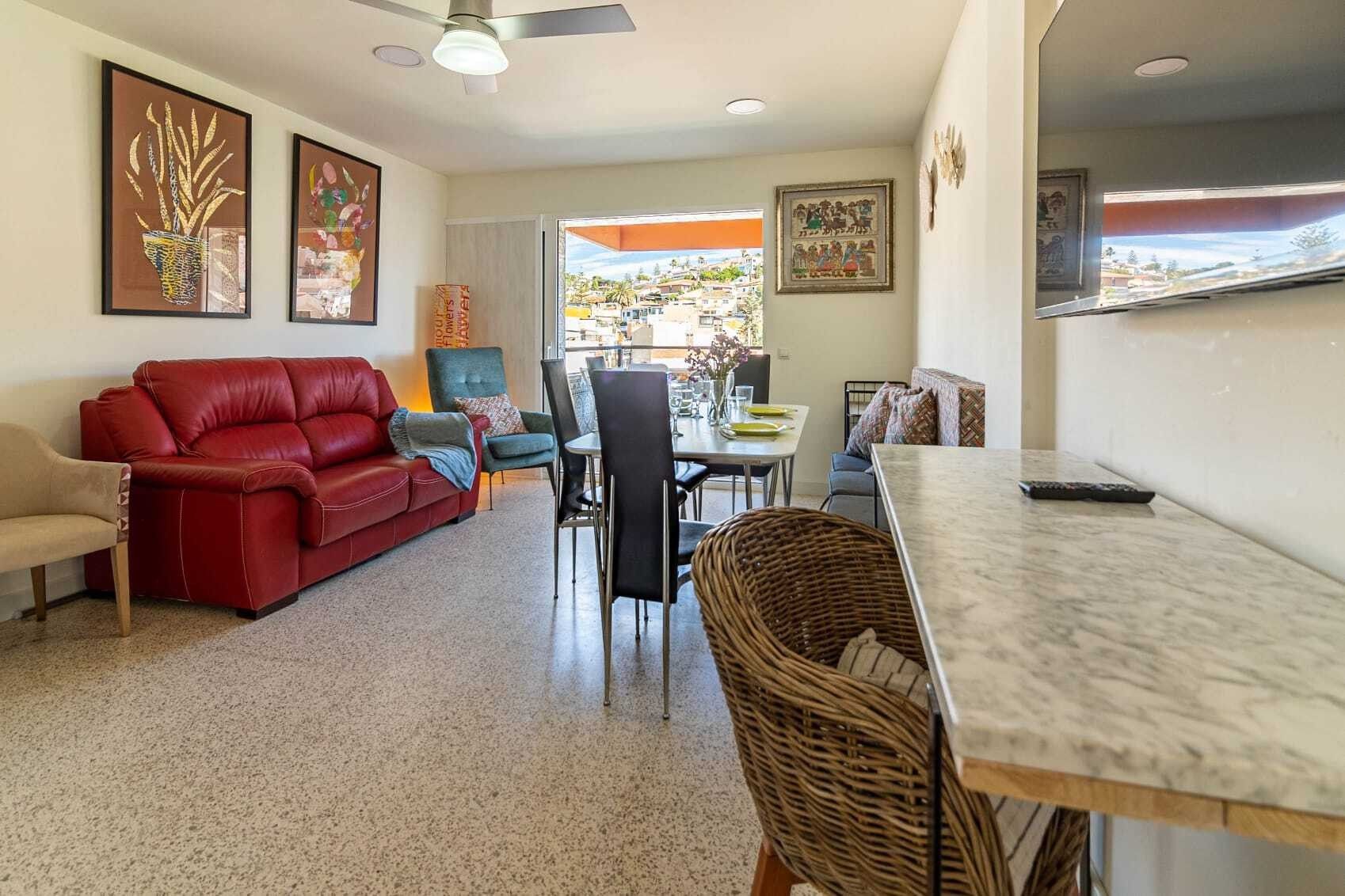 Una sala de estar con un sofá rojo, una mesa de comedor con sillas y una cocina visible a través de una puerta abierta.
