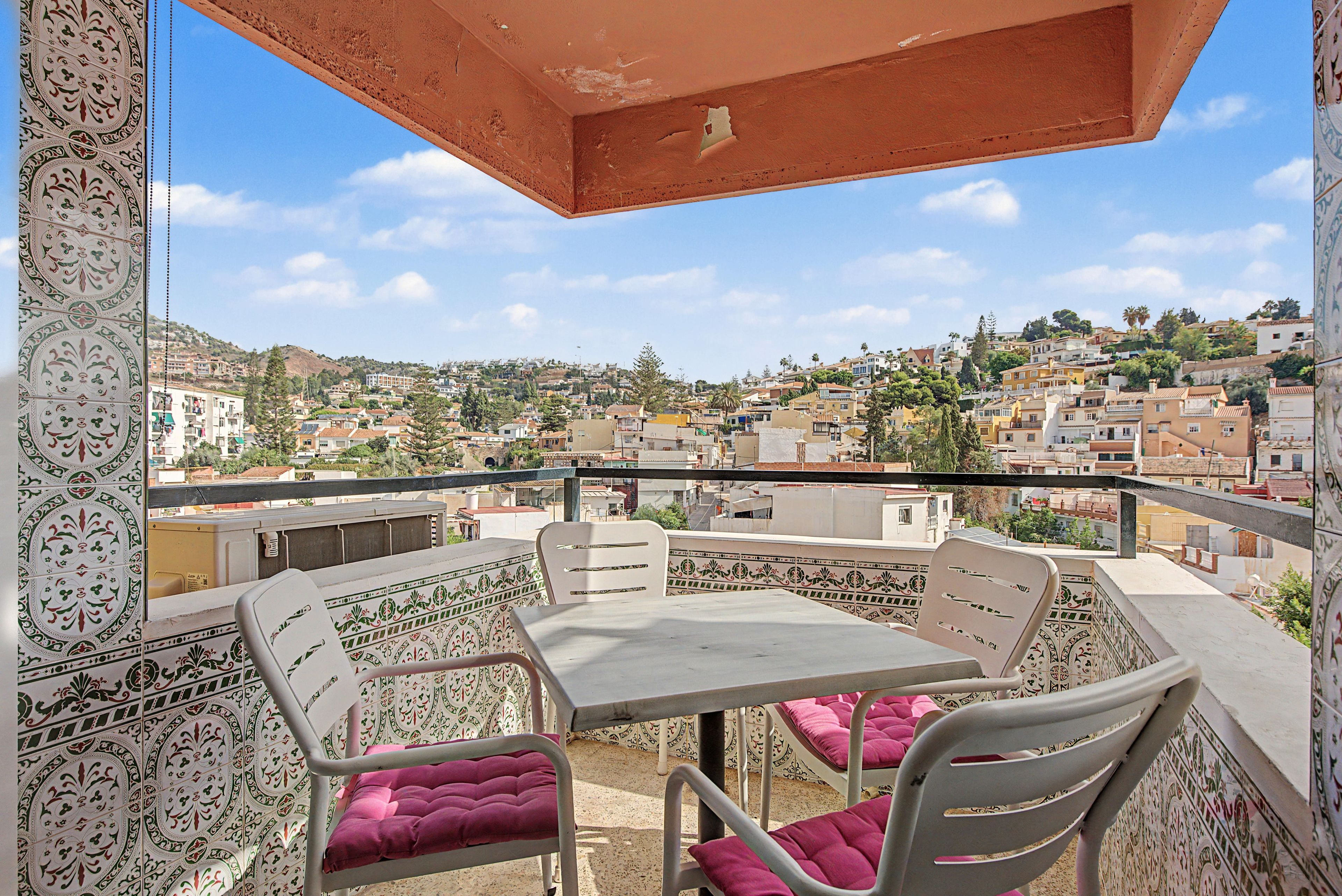Un balcón con mesa y sillas, con vistas a un pueblo en una ladera densamente poblada.