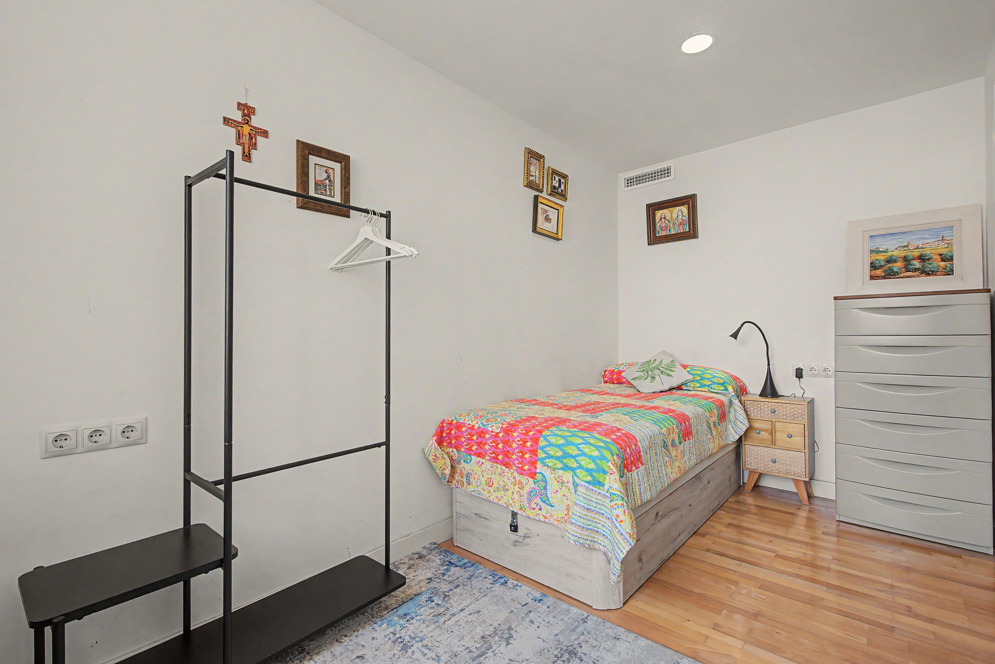 Un dormitorio con cama, armario, banco, radiador y una pared con cuadros.