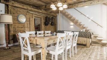 A rustic wooden interior with a dining table, white chairs, a chandelier, a clock, and a staircase.