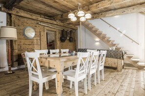 A rustic wooden interior with a dining table, white chairs, a chandelier, a clock, and a staircase.