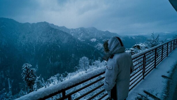 Eine Person steht auf einem mit Schnee bedeckten Balkon und blickt auf eine gebirgige Landschaft.