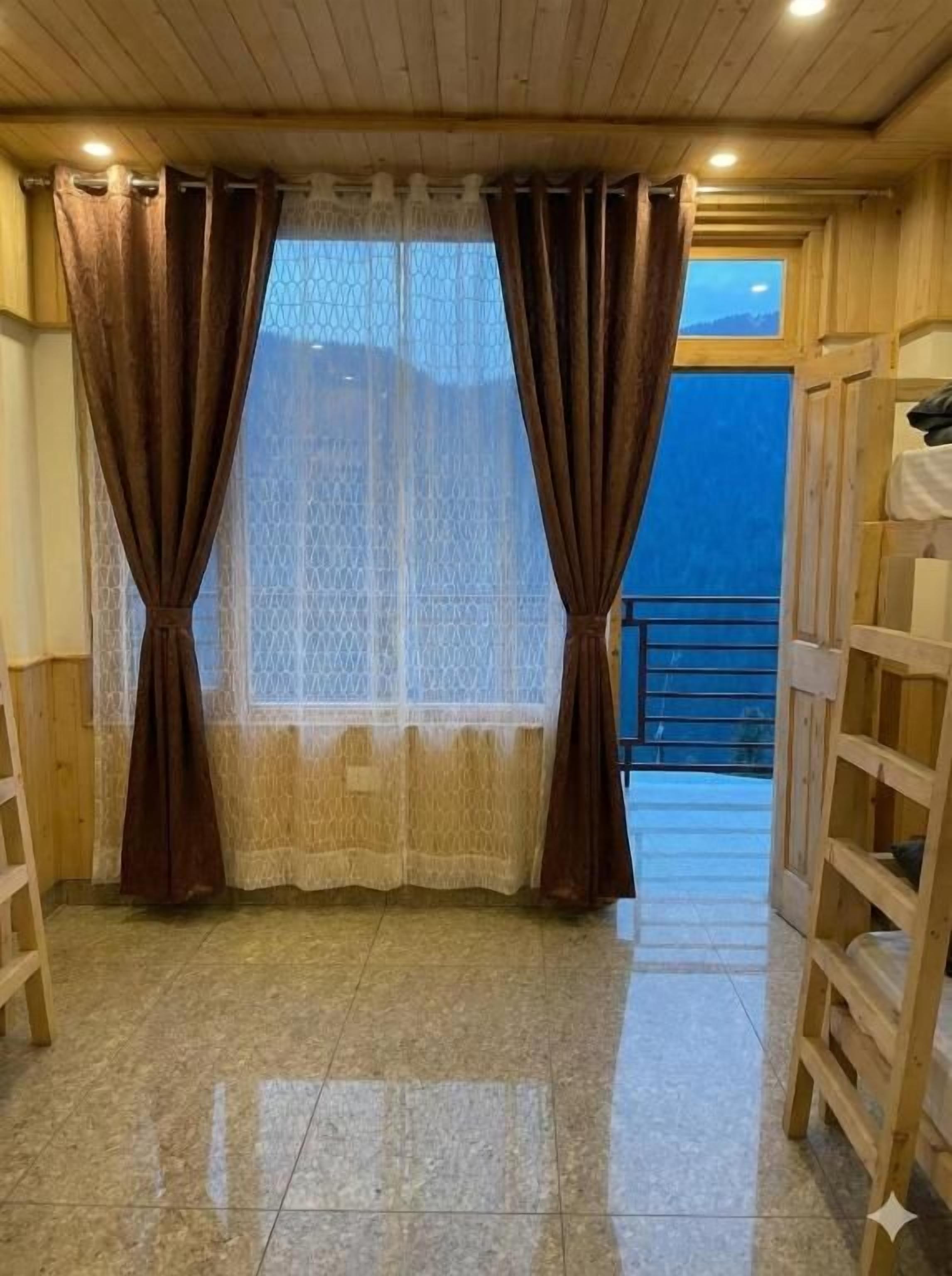 A room with a wooden floor, a window with brown curtains, and a view of a balcony.