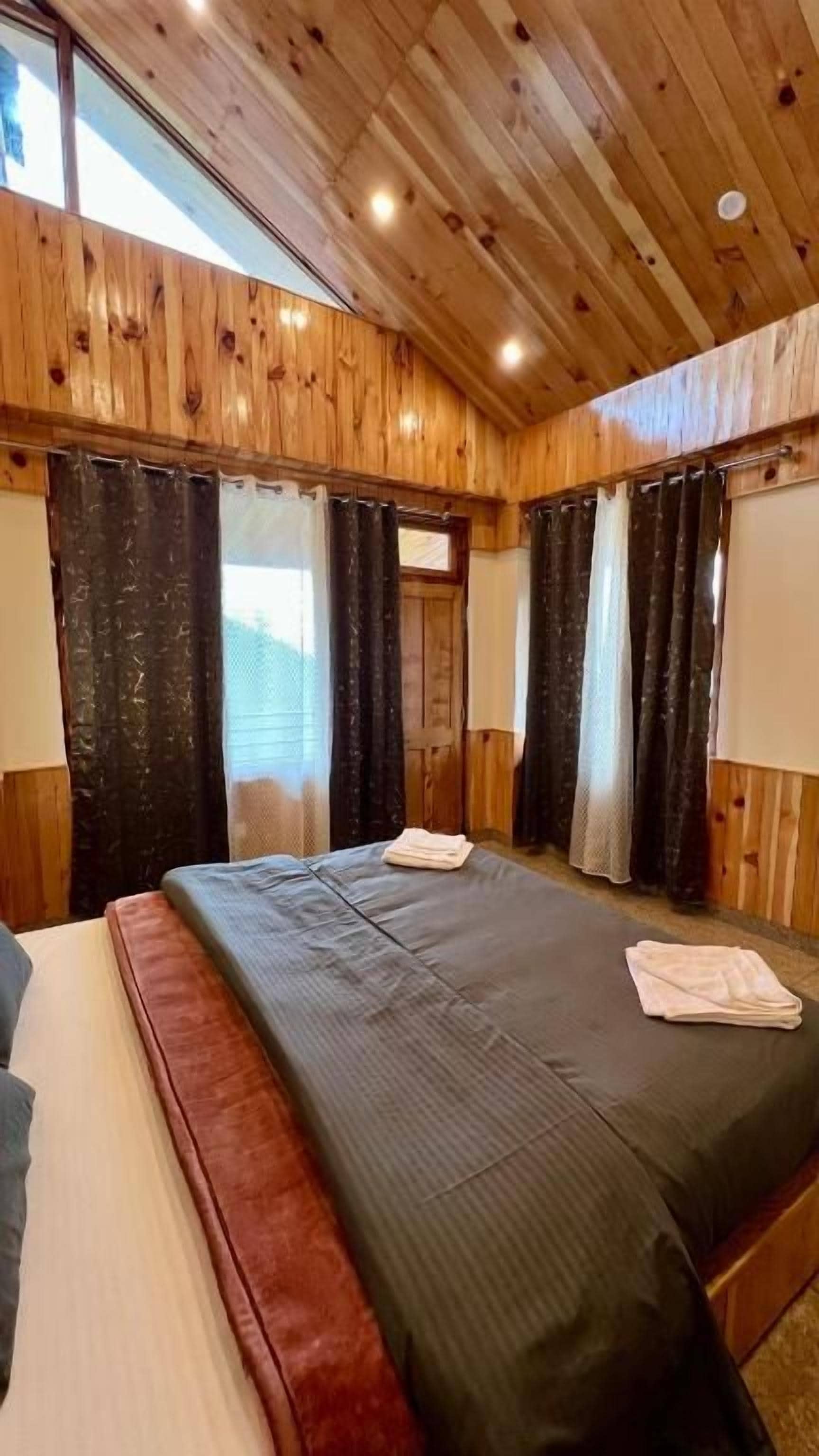 A wooden-paneled room with a large bed, two windows with curtains, and a wooden ceiling.