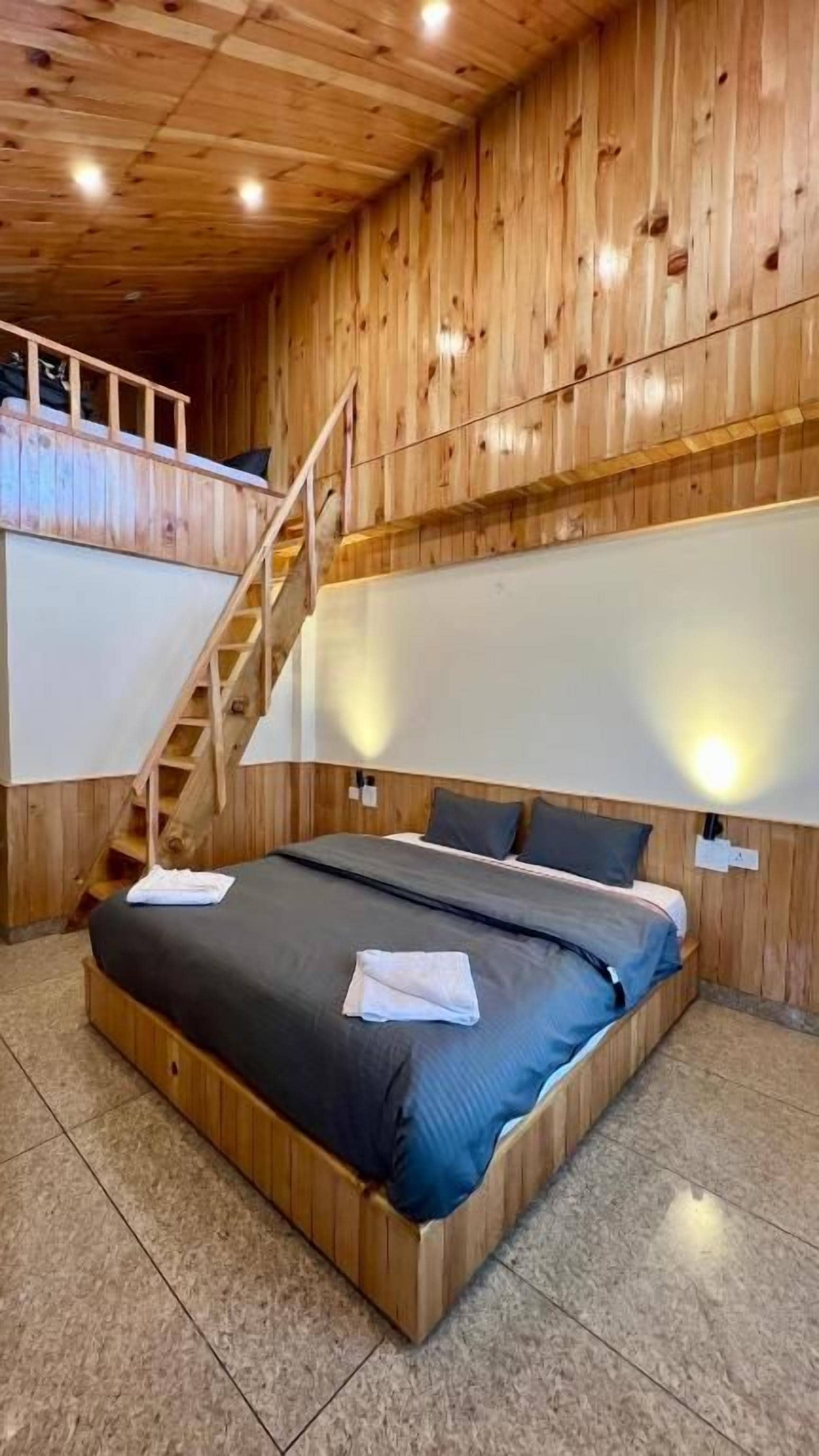 A wooden-paneled room with a bed, a staircase, and two wall-mounted lights.