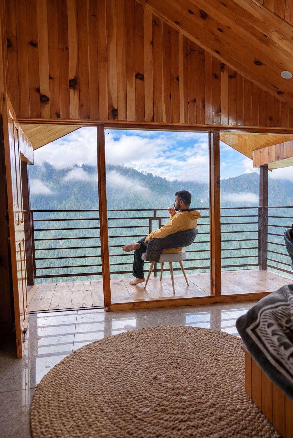 Una persona sentada en un taburete frente a un amplio ventanal con vistas a un paisaje montañoso.