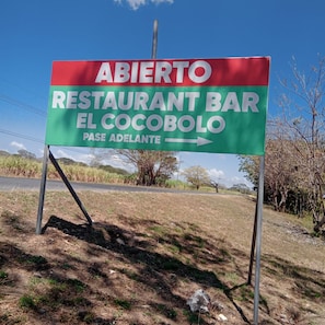 Un panneau indicateur pour un restaurant-bar appelé « ABIERTO RESTAURANT BAR EL COCOBOLO », avec une flèche pointant vers la droite.