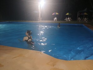 A swimming pool with two people in the water, surrounded by a sandy area and a few umbrellas. - www.elcocobolofoodanrest.com (Canas Dulces)