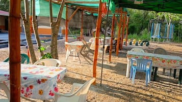 Un espacio de estar al aire libre con mesas y sillas, cubierto por toldos verdes y rodeado de árboles.
