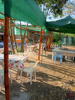 Un espacio de estar al aire libre con mesas y sillas, cubierto por toldos verdes y rodeado de árboles.