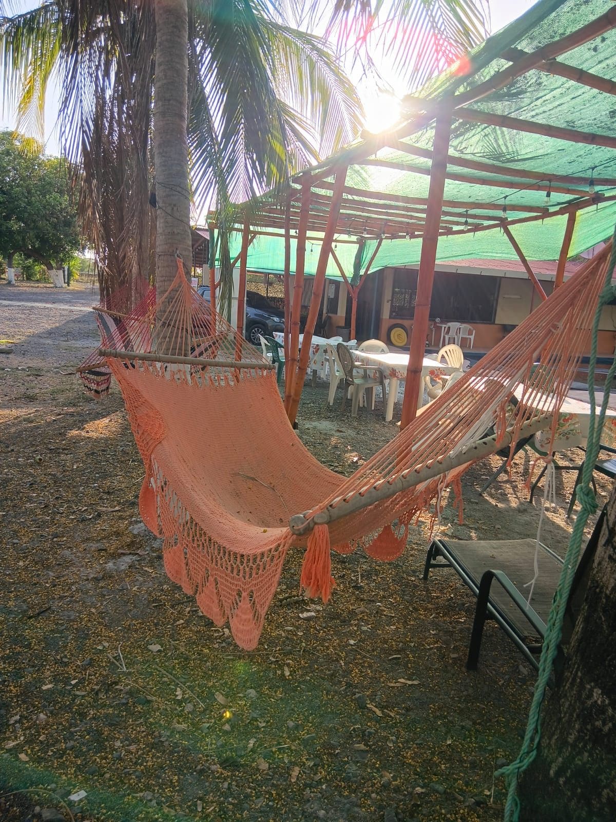 A hammock under a shaded structure, surrounded by plastic chairs and tables, with a building in the background.