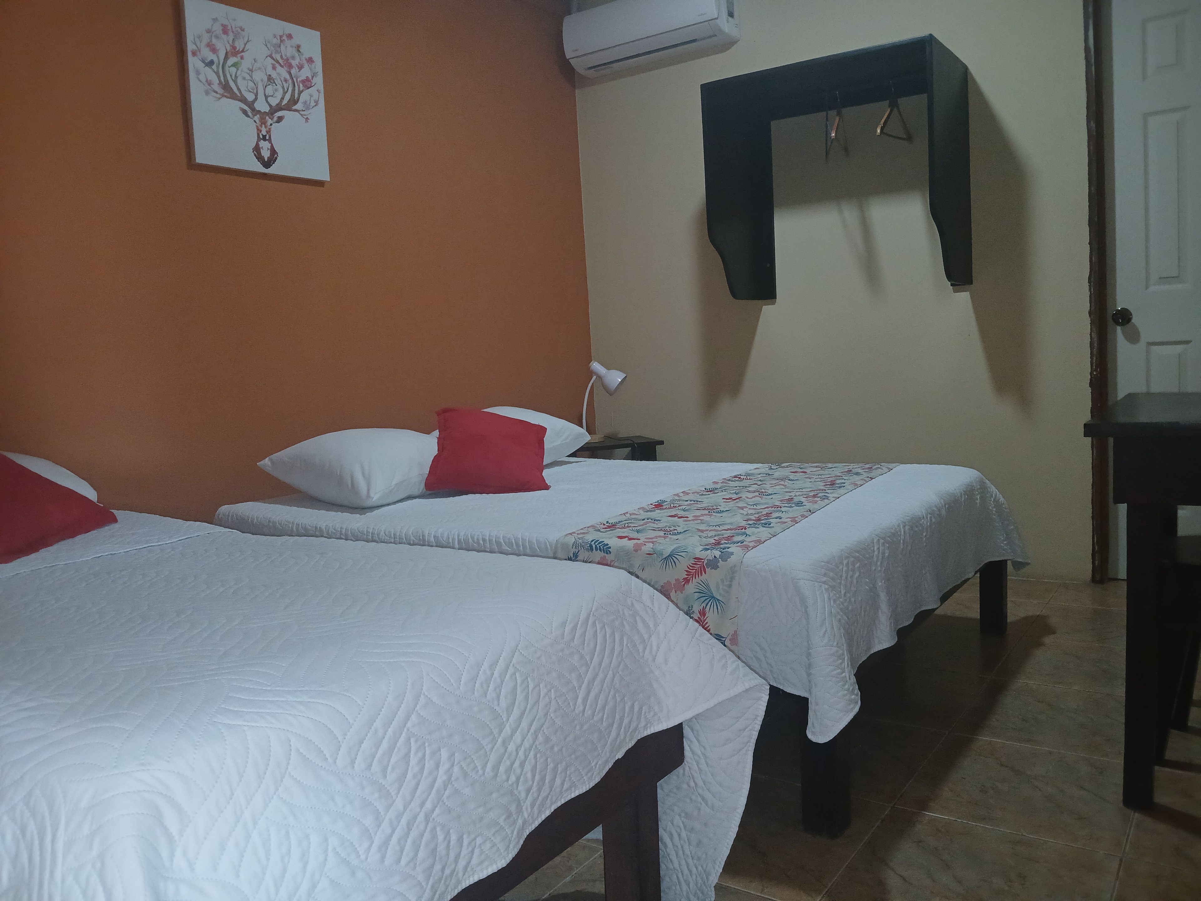 Habitación de hotel con dos camas, un almohadón rojo, un almohadón blanco, aire acondicionado de pared, una lámpara de noche y una puerta.