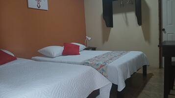 Habitación de hotel con dos camas, un almohadón rojo, un almohadón blanco, aire acondicionado de pared, una lámpara de noche y una puerta.