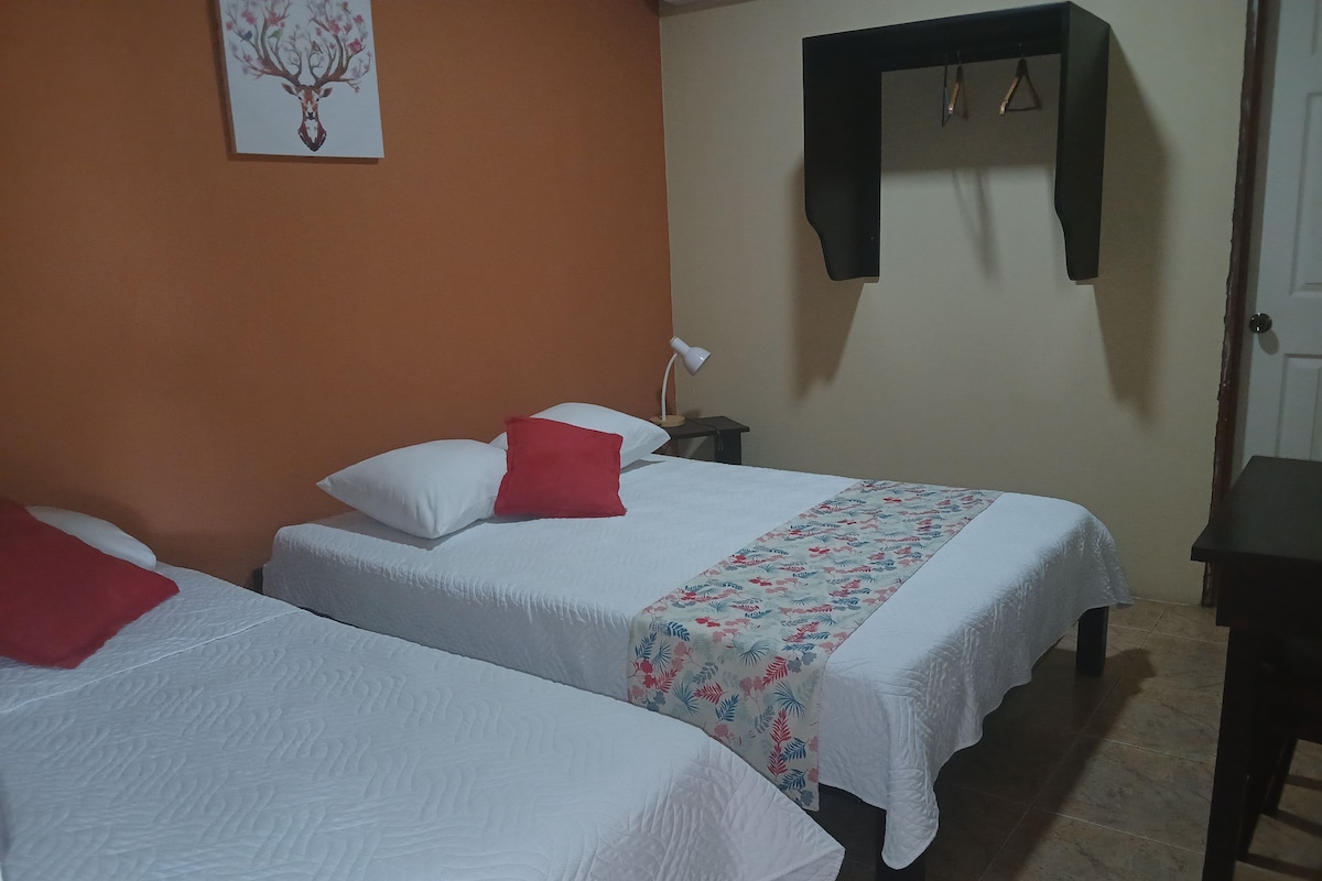 Una habitación de hotel con dos camas, cada una con ropa de cama blanca y almohadas rojas, una mesita de noche con lámpara y un estante en la pared.