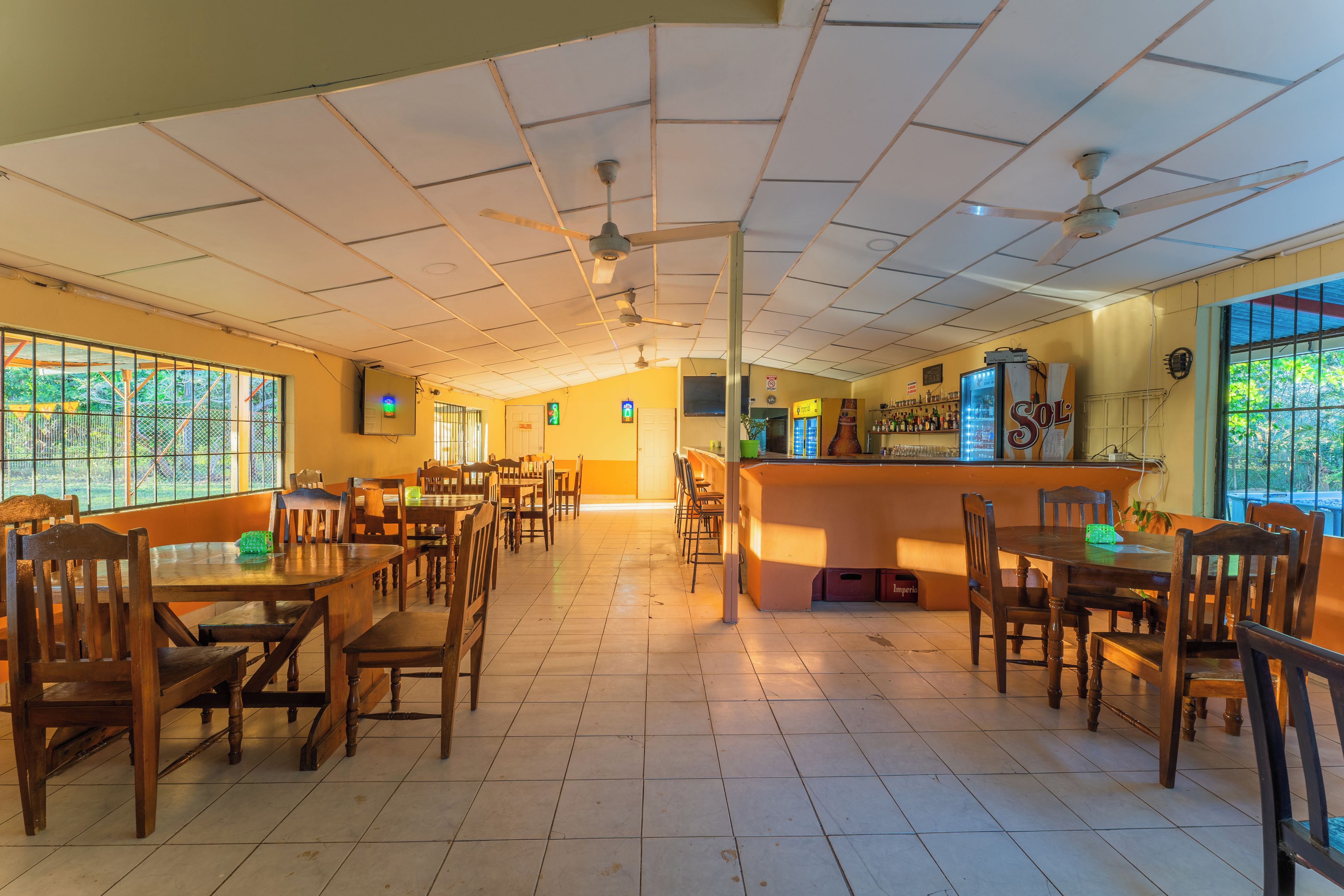 A restaurant with multiple wooden tables and chairs, a bar area, and a ceiling with fans.