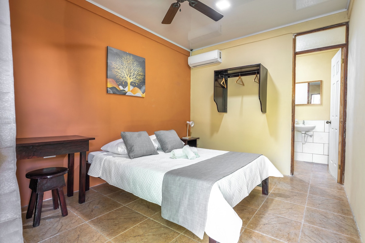 Un dormitorio con cama, escritorio, silla, un cuadro, equipo de aire acondicionado y un baño visible a través de una puerta abierta.
