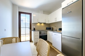 A compact kitchen with white cabinets, a stainless steel refrigerator, a microwave, and a stove. There is a dining table with chairs and a view through a glass door. - Villa in Lanzarote | Pool | Volcanic Garden (Yaiza)