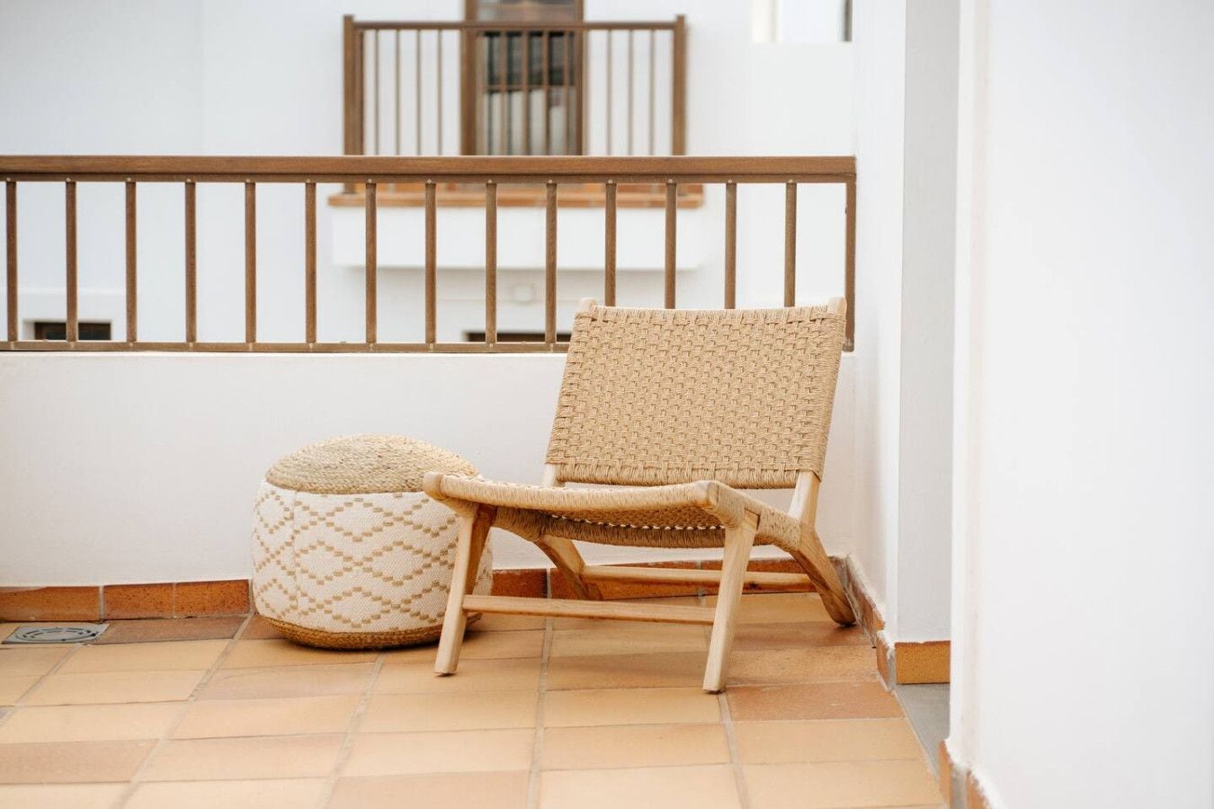 A wicker chair with a matching ottoman on a tiled floor, a wooden railing, and a white wall in the background.