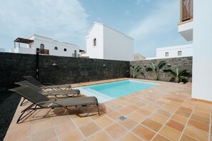 A swimming pool with lounge chairs, a stone wall, and white buildings in the background. - Villa in Lanzarote | Pool | Private Garden (Yaiza)