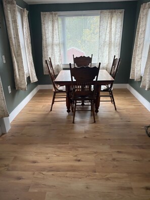 Uma área de refeições com mesa e cadeiras de madeira, janela com persianas e parede verde.