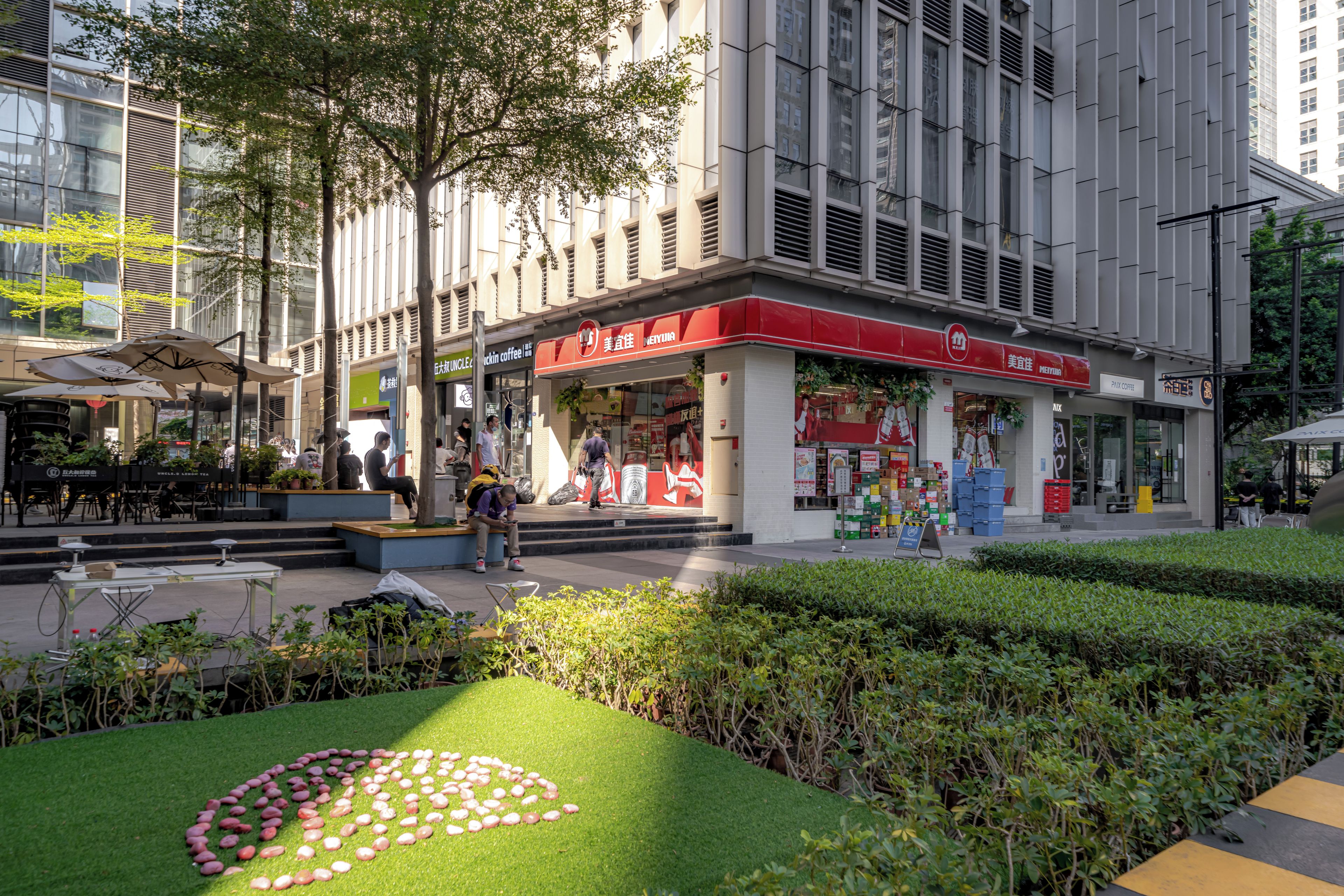 Una calle urbana moderna con una cafetería, asientos al aire libre y una zona ajardinada con un círculo de piedras.