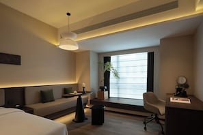 Una camera d'albergo moderna con un letto, un divano, una scrivania, una sedia, una pianta in vaso e un'ampia finestra.