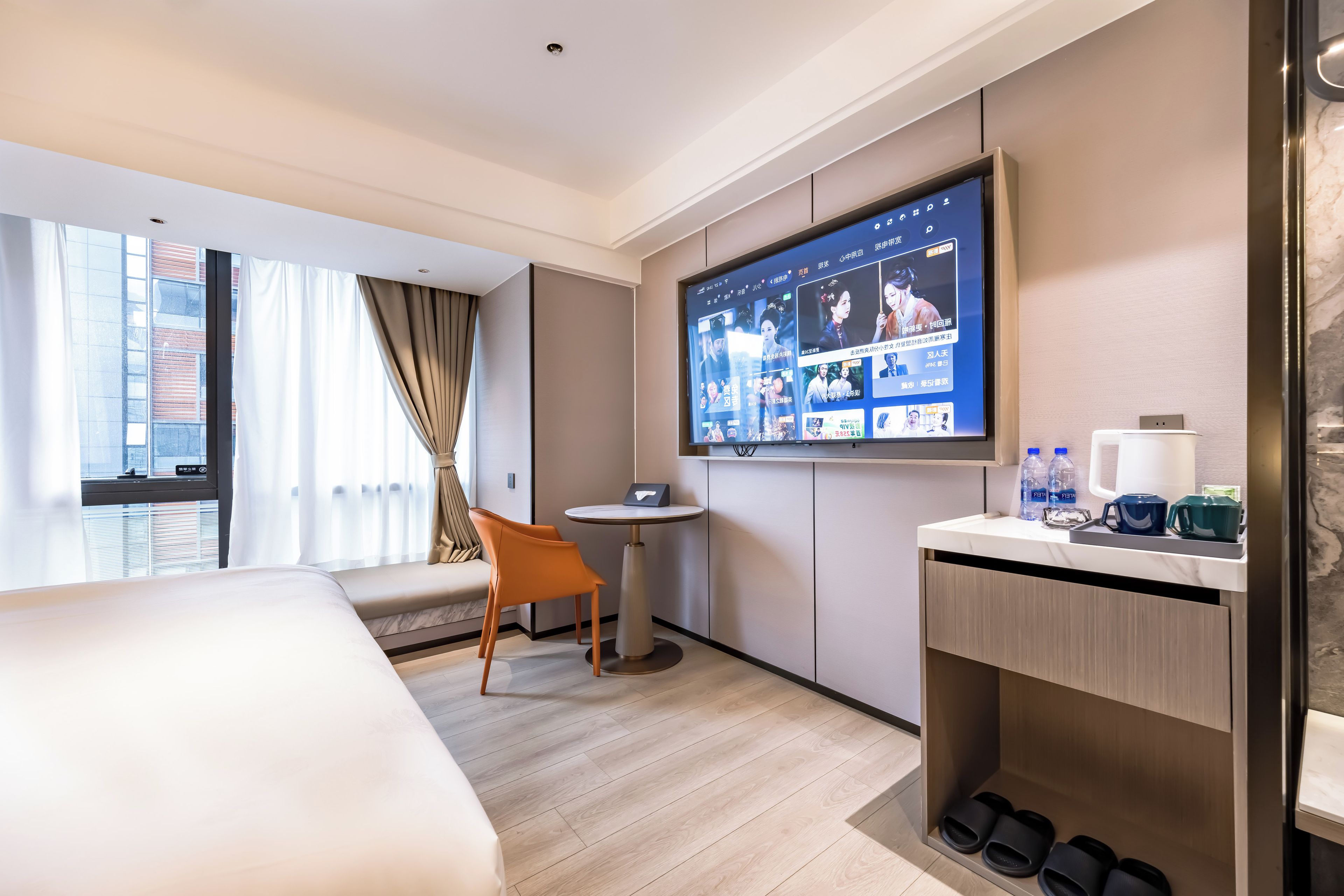 A hotel room with a bed, a TV mounted on the wall, a small round table, a chair, and a shoe rack.