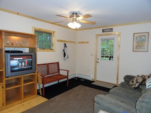 A living room with a television, a wooden bench, a couch, a ceiling fan, and a door labeled 'CABINS RENTAL'. - "Inn The Pines" 2-bedroom apartment in charming Jay, NY (Jay)
