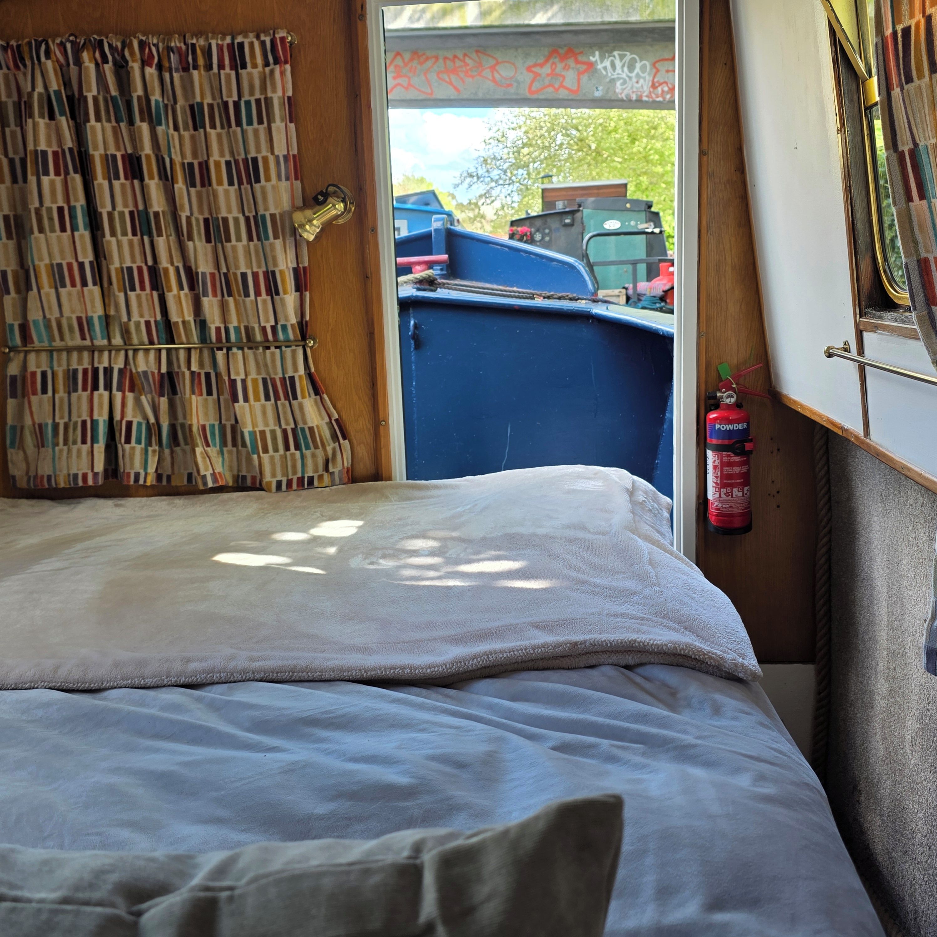 A bed with a white blanket, a window with a view of parked vehicles, and a fire extinguisher on the wall.