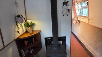 A room with a wood-burning stove, a shelf with books and a plant, and a window with a view of the outside.
