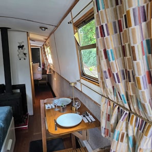 Et spiseområde med et bord dækket til to, et vindue med gardiner og et tekøkken i baggrunden.