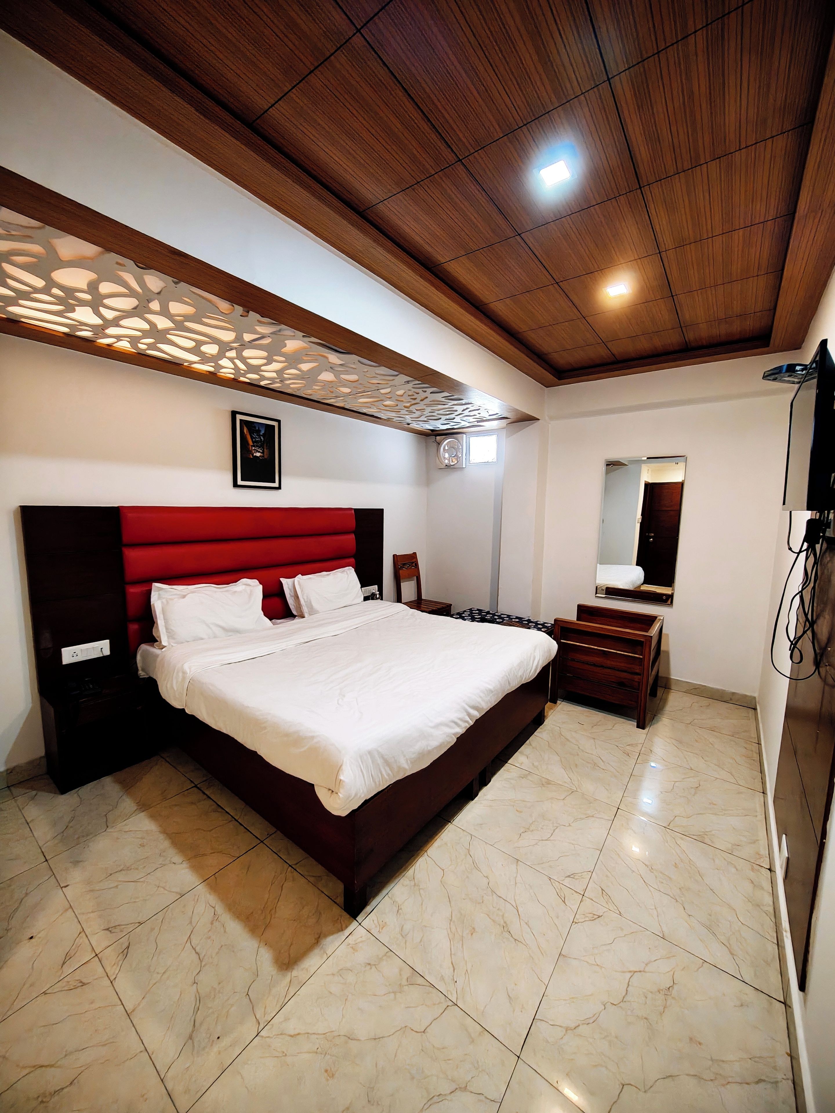 Habitación de hotel con una cama grande, cabecera de madera, mesita de noche, espejo y televisor.