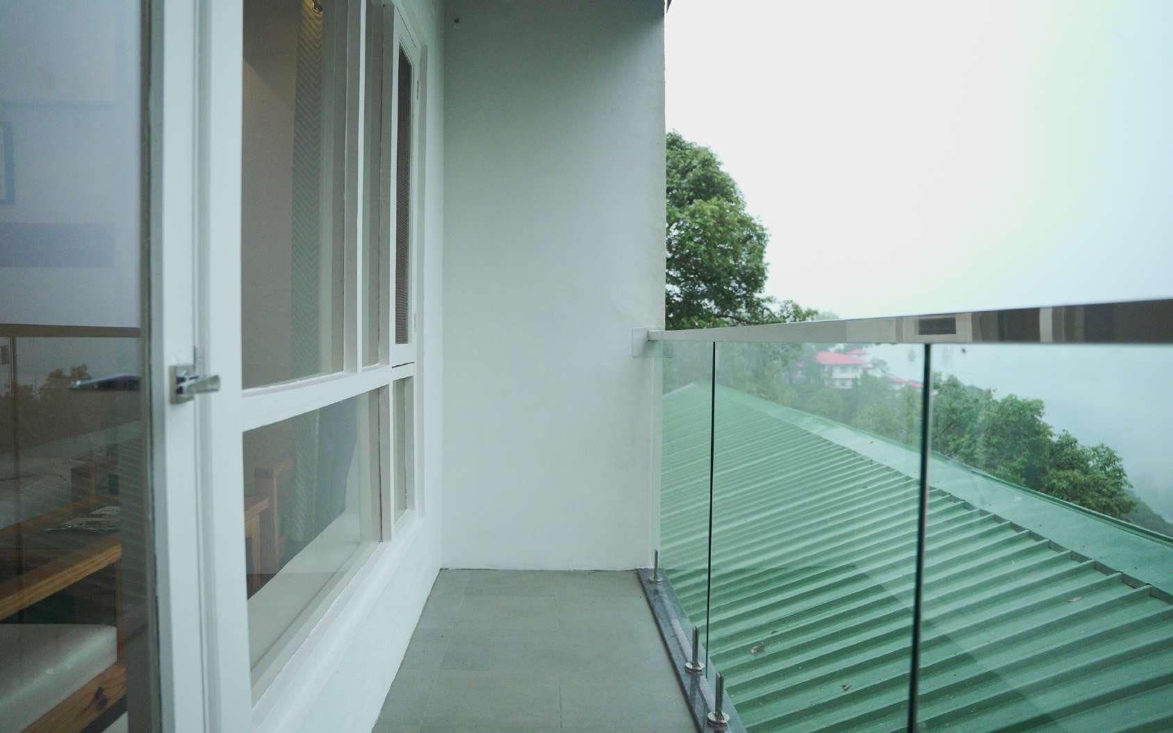 Un balcón con barandilla de vidrio, una puerta blanca y vistas a un techo verde y árboles.
