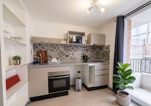 Cocina compacta con microondas, horno y lavamanos. Hay repisas con frascos y una planta en maceta.