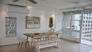 Un comedor moderno con una mesa blanca, sillas y un banco. Hay una pared con una lista de normas de la casa y un cuadro sobre la mesa.