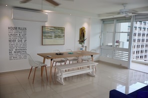 Un comedor moderno con una mesa blanca, sillas y un banco. Hay una pared con una lista de normas de la casa y un cuadro sobre la mesa.