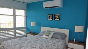 Un dormitorio con una cama, dos mesitas de noche, aire acondicionado y una ventana con persianas.