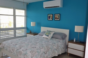 Un dormitorio con una cama, dos mesitas de noche, aire acondicionado y una ventana con persianas.