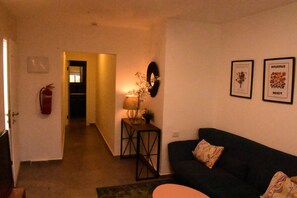 Una sala de estar con un sofá oscuro, una mesita auxiliar con una lámpara y dos cuadros en la pared.