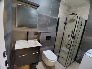 Un baño moderno con lavamanos blanco, muebles oscuros, inodoro y cabina de ducha con paredes de vidrio.