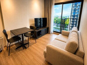 A modern living room with a sofa, a dining table with chairs, a TV on a stand, and a large window with a city view. - 1Br Condo for Long Stay in Ekkamairamindra (Bangkok)