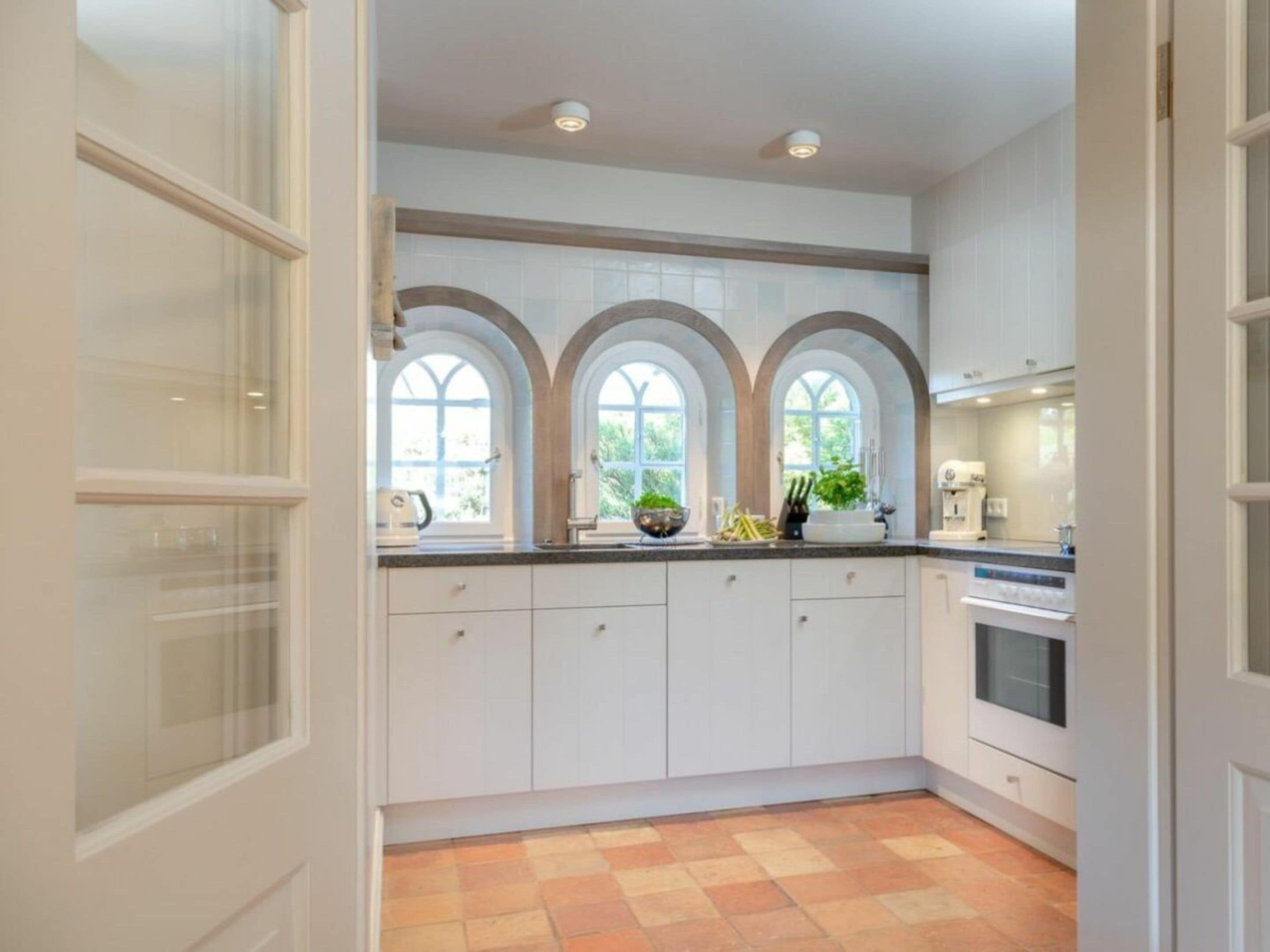 A kitchen with white cabinets, a tiled backsplash, and three arched windows.