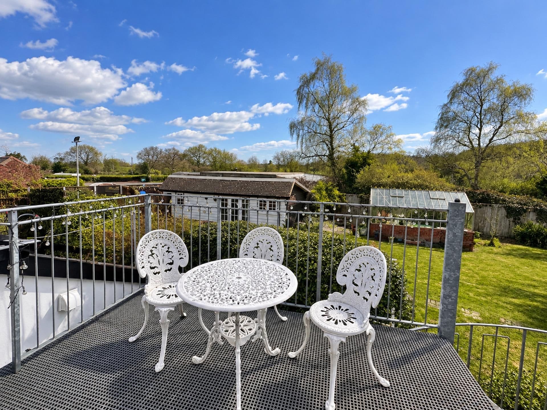 A patio with a metal table and chairs, surrounded by a metal fence, with a view of a house and trees in the background.